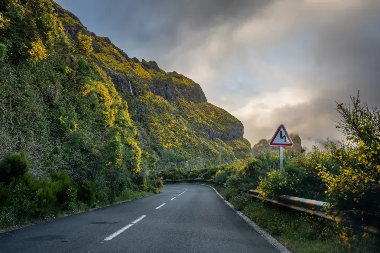 A mountain road below a rock face, with a hazard sign in front