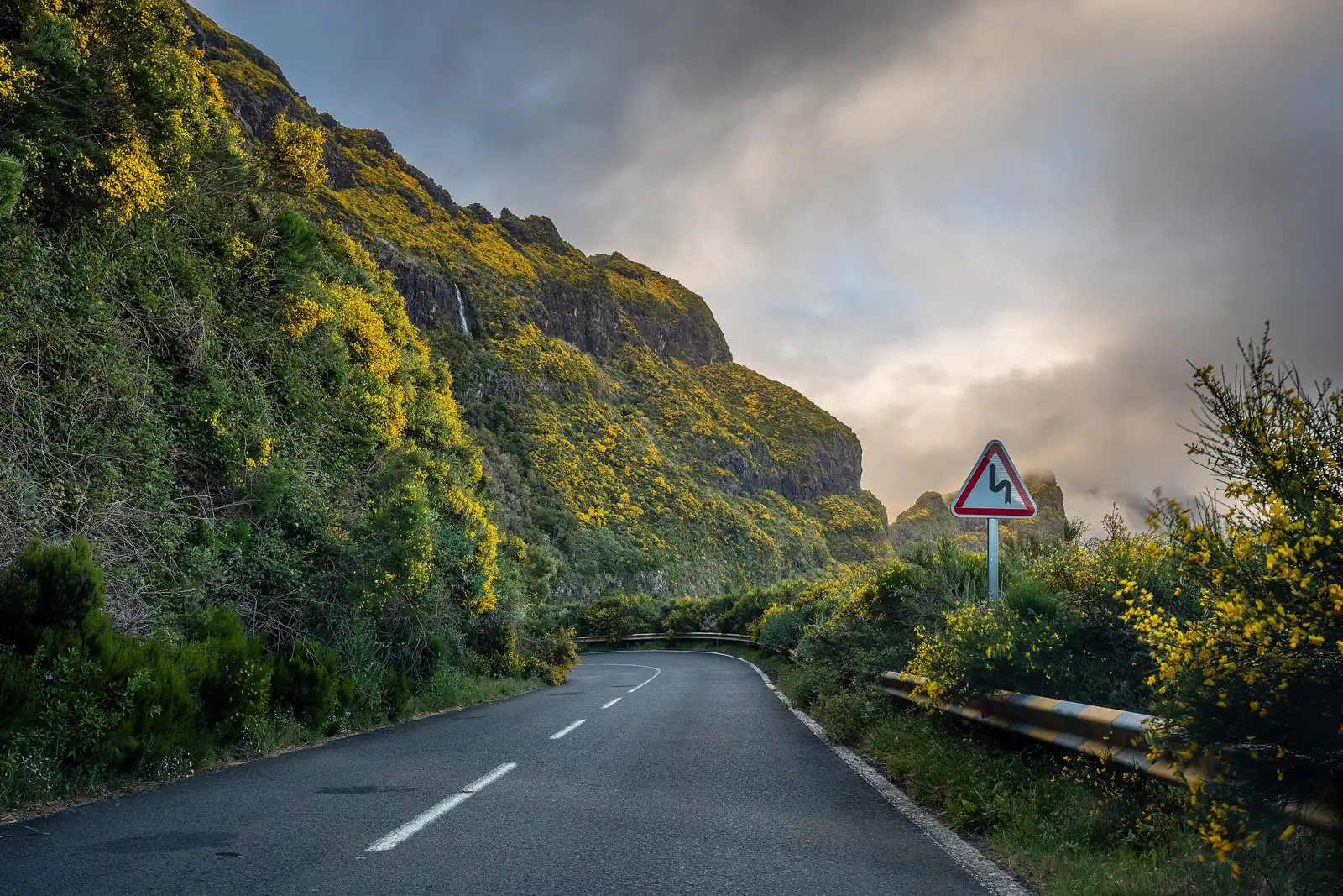 A mountain road below a rock face, with a hazard sign in front